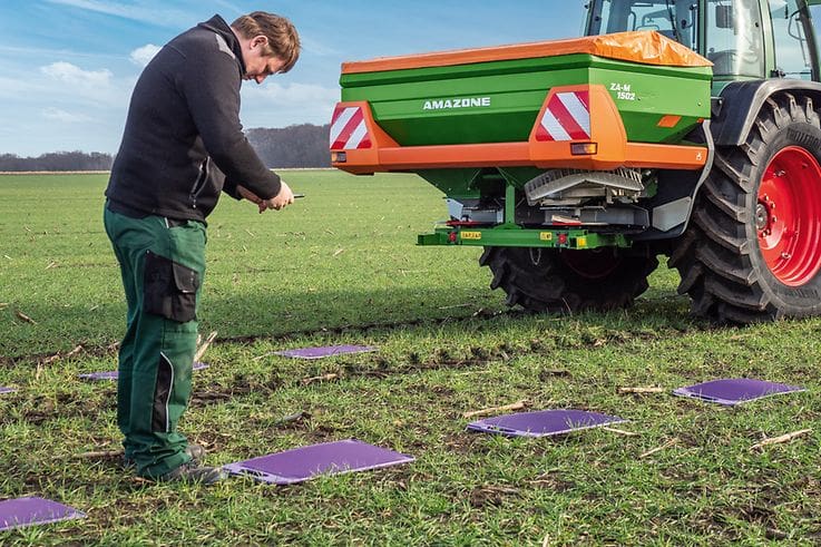 Man standing in field using EasyCheck system with AMAZONE ZA-M mounted spreader Infront of him